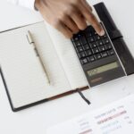 A hand using a calculator with financial documents and notebook, depicting an office setup for financial analysis.